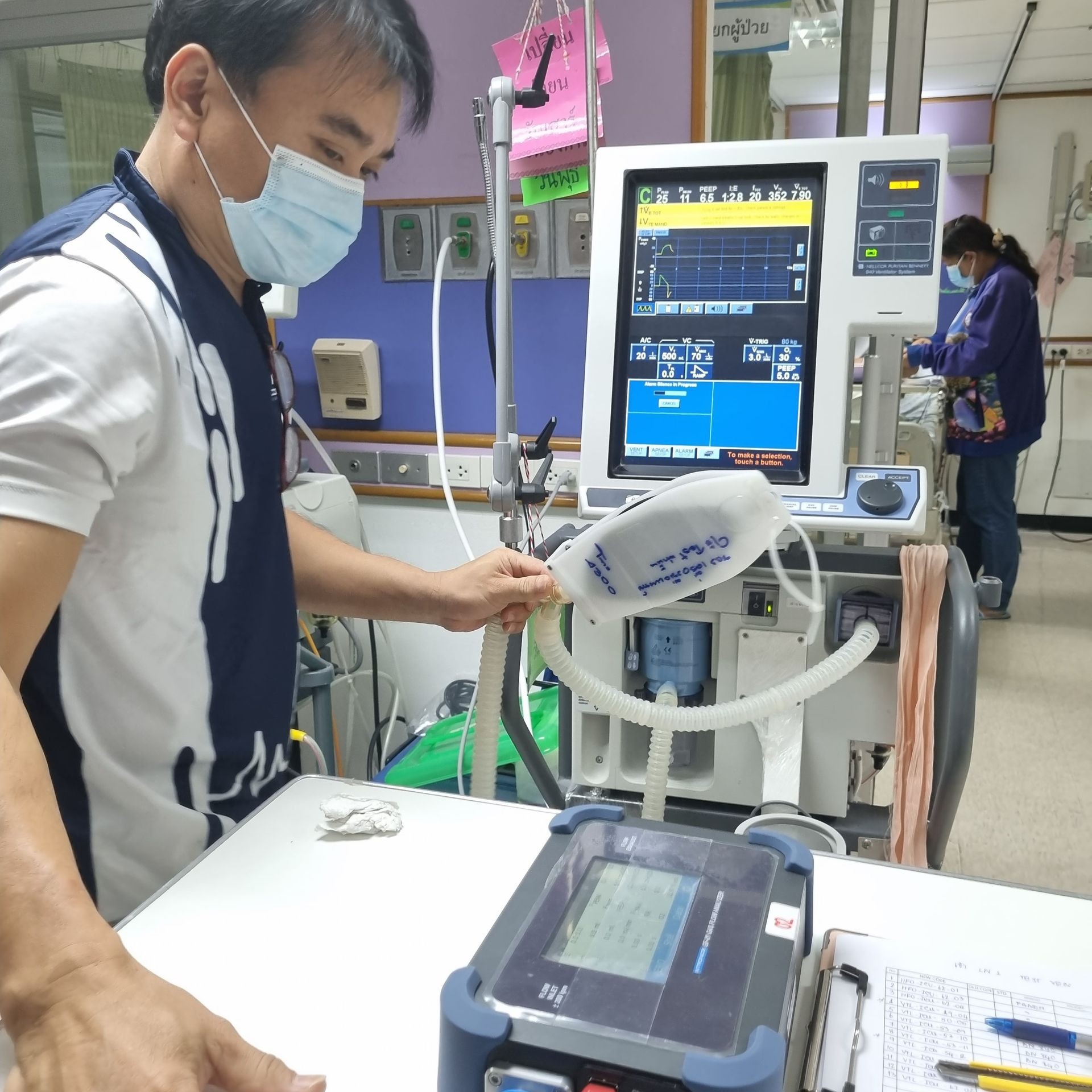 Man wearing a mask operating medical ventilator equipment, with monitors and tubing in a hospital setting.