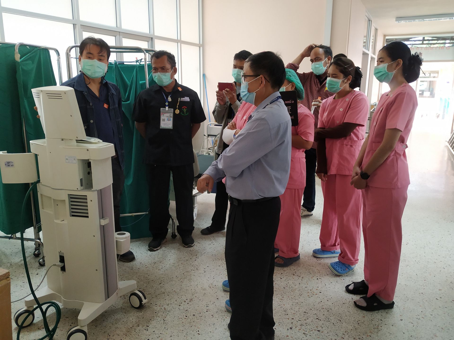 Medical professionals in masks gather around a machine in a hospital hallway for a demonstration or training.