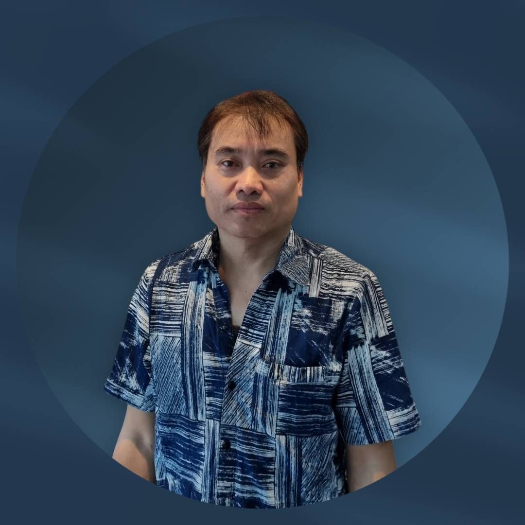 Man in a patterned blue shirt standing against a dark blue background, looking at the camera.